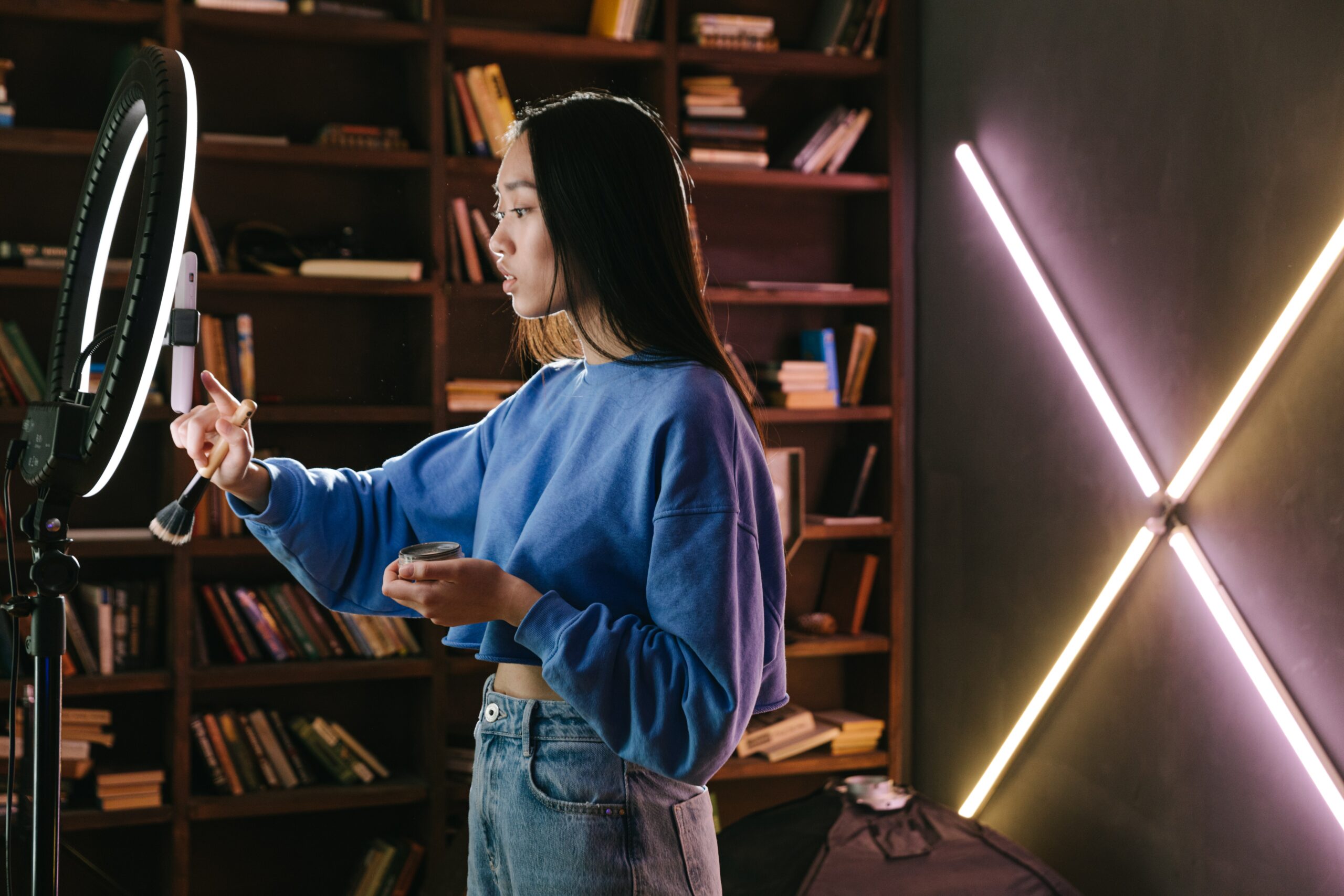 A young woman in profile standing in front of a ring light and making a TikTok on her phone